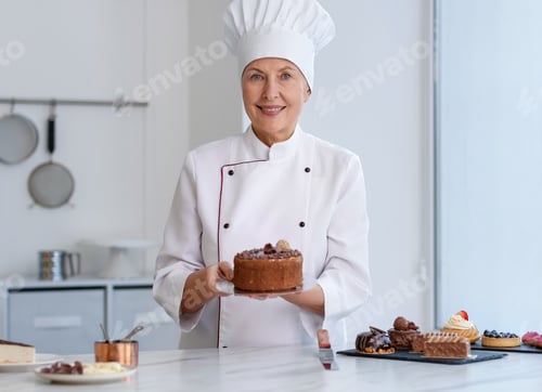 Preview: Smiling pastry chef holding tasty cake at table with other desserts in kitchen