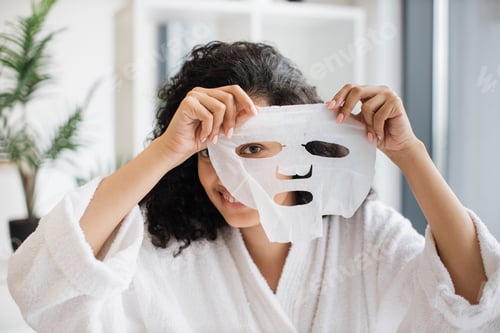 Preview: Female in white bathrobe looking through holes of cosmetic sheet mask