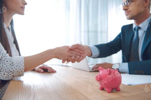 Preview: selective focus of insurance agent and client shaking hands at tabletop with pink piggy bank