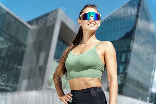 Preview: Confident female runner posing with hands on hips, wearing reflective sunglasses, cityscape behind.
