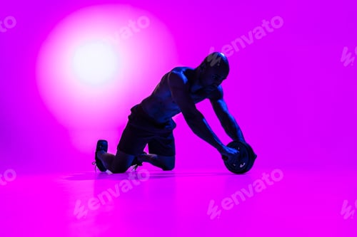 Preview: Bodybuilder working on his abs with a ab roller on studio background with color filter