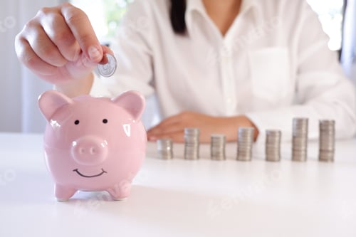 Preview: Businessperson Inserting Coins In Piggy Bank With Stack Of Coins Over The Desk.