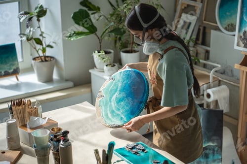 Preview: Craftswoman in respirator inspect dried epoxy resin painting with waves, checking result in workshop