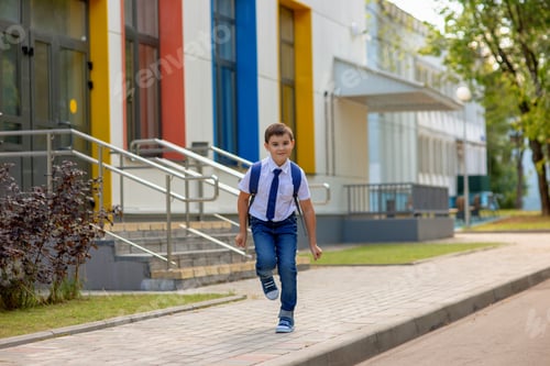 Preview: Cheerful young schoolboy in white shirt, blue tie and backpack runs from school