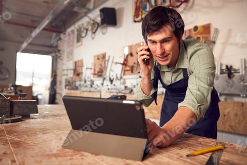 Preview: Male Business Owner In Workshop Using Digital Tablet And Making Call On Mobile Phone