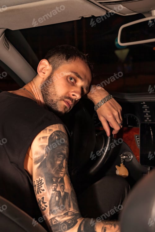 Preview: Young man driver sitting at steering wheel of car at night thoughtfully looking at camera