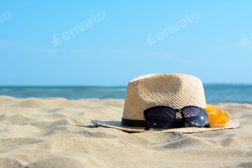 Preview: Hat with beautiful sunglasses and flowers on sand near sea. Space for text