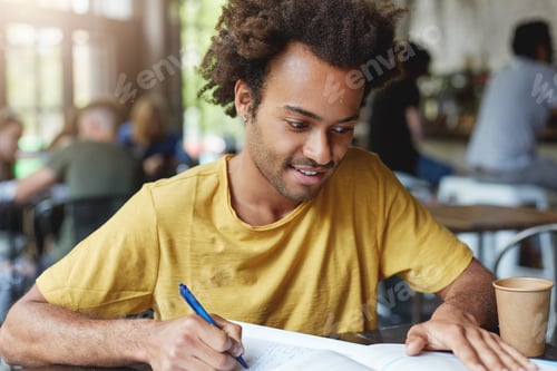 Preview: Focused student male with dark bushy hair and bristle wearing casual T-shirt writing something in hi