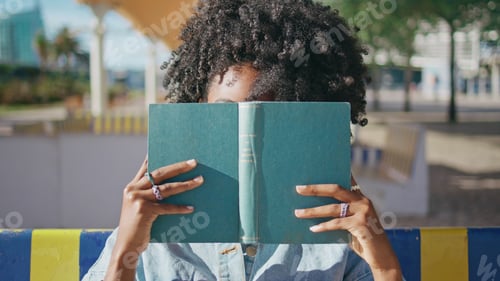 Preview: Young girl peeping book sitting at sunny street close up. African girl posing