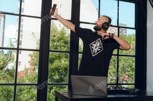 Preview: young male dj with Arab features taking a selfie with his phone while working mixing music.