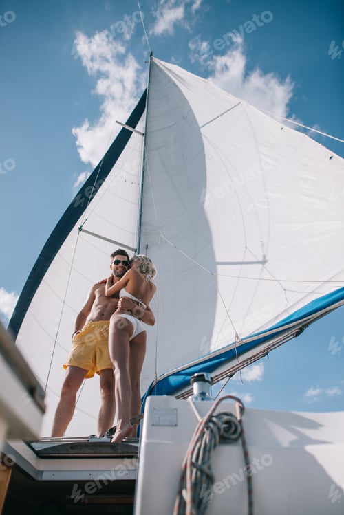 Preview: low angle view of shirtless man in sunglasses embracing girlfriend in bikini on yacht