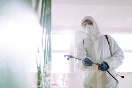 Preview: Man in Hazmat suit, protective gloves and goggles use sprayer equipment disinfect the entrance.