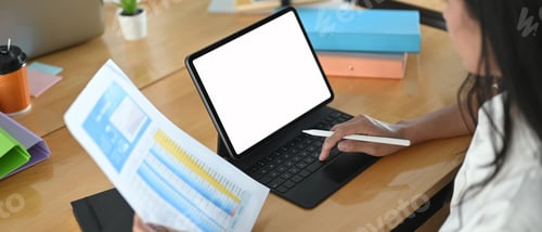 Preview: A woman is using a computer tablet with a white blank screen at the wooden working desk.