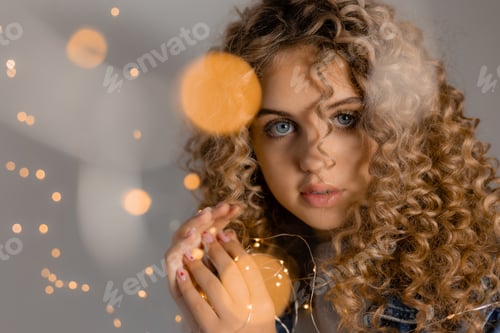 Preview: portrait of a young woman with blue eyes and curls holding glowing lights of a garland in her hands