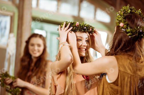 Preview: Beautiful young women in boho style wearing floral wreaths and looking at each other