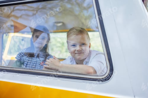 Preview: Cute happy boy and girl look out of the window of a yellow and white retro minivan.