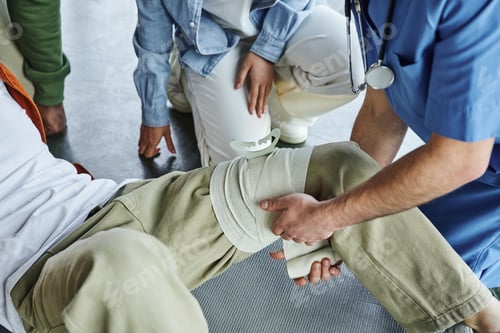 Preview: partial view of professional paramedic applying compressive bandage on leg of man near multiethnic