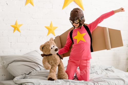 Preview: Kid with cardboard wings standing on bed and holding teddy bear