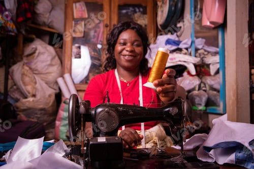 Preview: an African seamstress holds a bobbin in her hand and works in her tailor shop,
