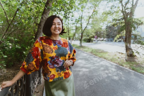 Preview: Stylish lady wearing a trendy, floral patterned top stands by park railing