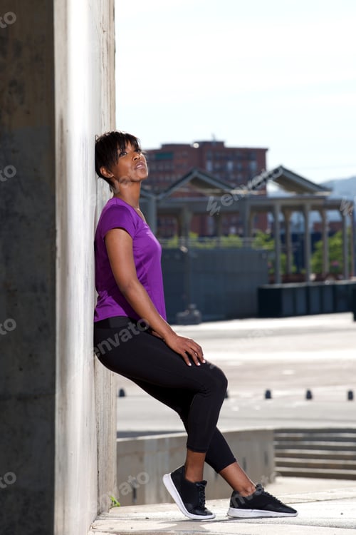Preview: Full length side of young african woman leaning to wall outdoors after workout
