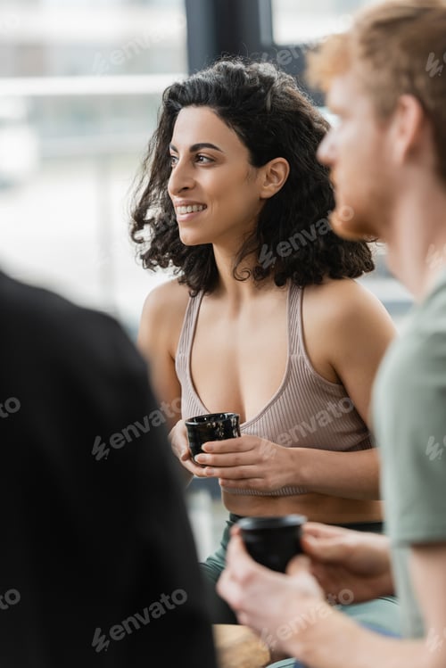 Preview: cheerful middle eastern woman with curly hair smiling during tea ceremony in yoga studio