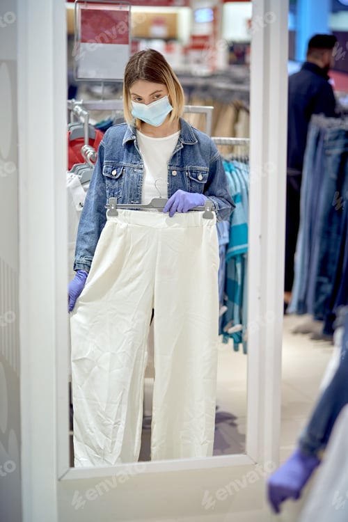 Preview: Young Blonde Girl Chooses Clothes Near The Mirror. Shopping During A Pandemic.