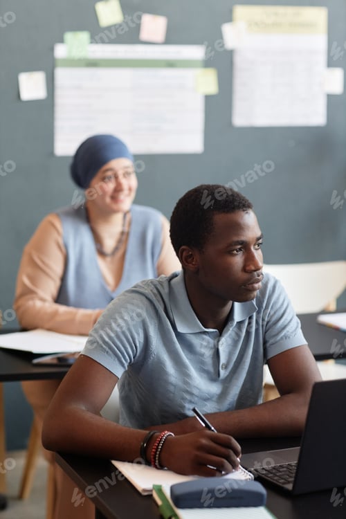 Preview: Young Black Man Writing in Notebook Listening to Teacher while Studying English in Classroom
