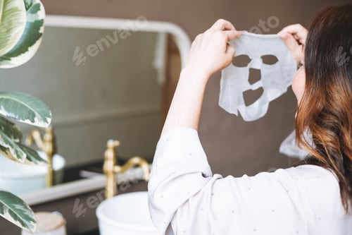 Preview: Adult brunette woman with sheet mask on her face in the bathroom at home