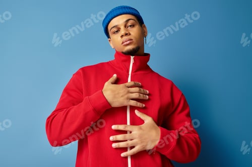 Preview: Trendy young man in a beanie and red jacket poses against a blue backdrop