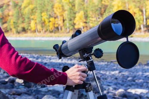 Preview: Traveler man on bank of mountain river tunes telescope to examine terrain and sky, selective focus.