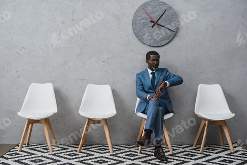 Preview: Handsome African american businessman sitting in a waiting room and looking at watch