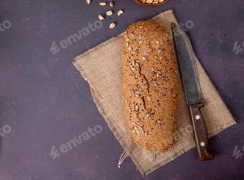 Preview: top view of seeded baguette and knife on sackcloth on right side and maroon background with copy