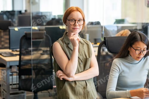 Preview: Young serious businesswoman in eyeglasses standing by desk next to her colleague