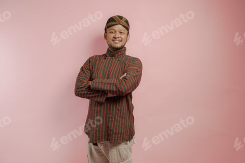 Preview: Confident smiling Asian man in a lurik and blangkon posing with arms folded.