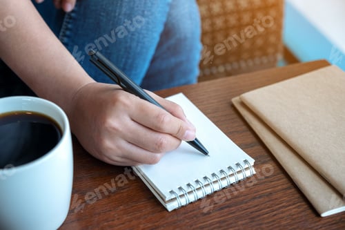 Preview: Closeup image of a woman's hand writing on blank notebook with coffee cup on wooden table