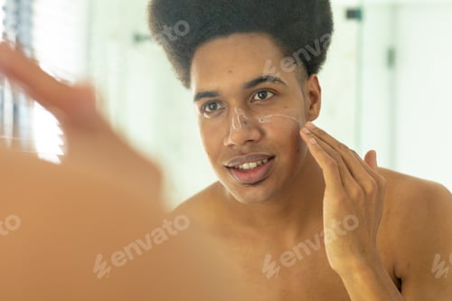 Preview: Happy biracial man looking in mirror and applying face cream in sunny bathroom