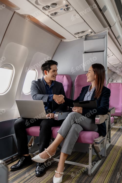 Preview: Asian businesspeople smiling and shaking hands while sitting on an airplane.