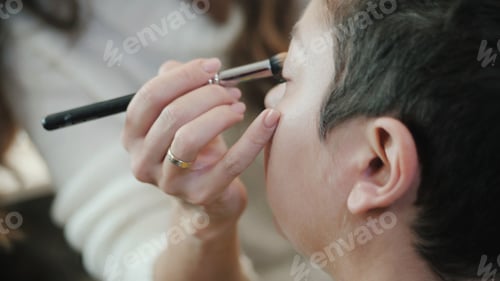 Preview: Esthetician - Closeup middle-aged woman having applied makeup by make-up artist in beauty salon.