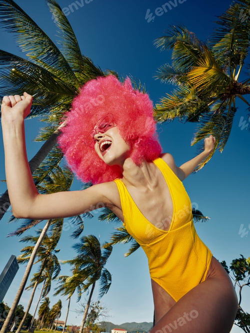 Preview: Woman in yellow swimsuit and pink wig, jumping with arms raised and smiling, vibrant and joyful