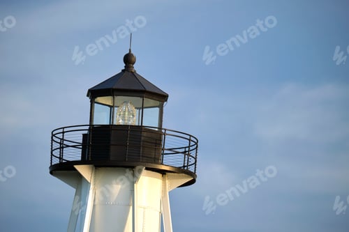 Preview: White tall lighthouse on sea shore against blue sky for commercial vessels navigation