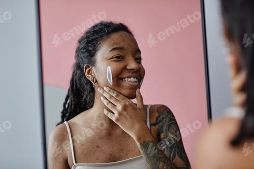 Preview: Candid smiling black woman with acne scars using face cream by mirror