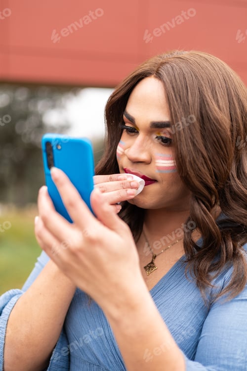 Preview: Young trans woman removing make up and using cellphone as mirror. LGTB lifestyle concept.