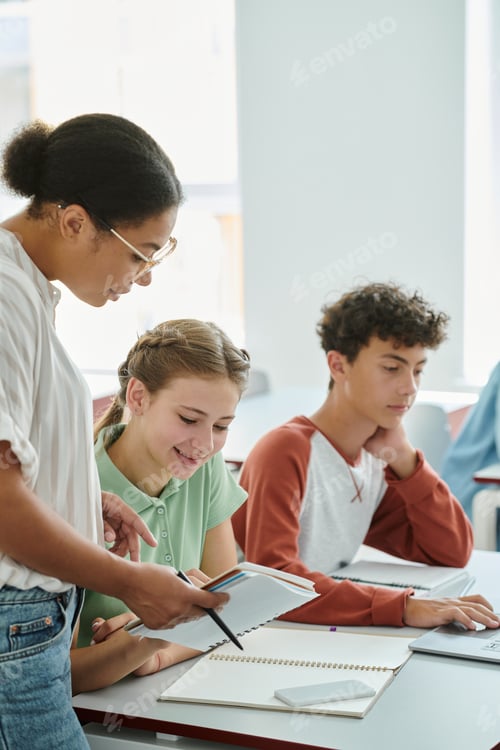 Preview: African american teacher holding notebook near positive schoolgirl during lesson in classroom