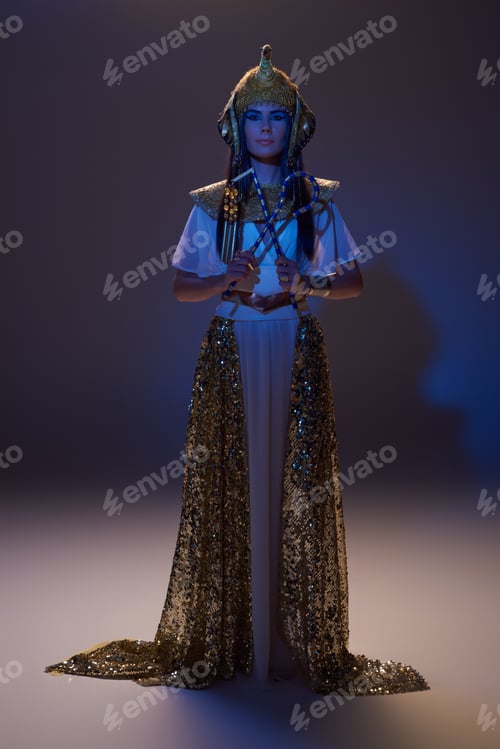 Preview: Full length of elegant woman in egyptian look holding crook and flail on brown with blue light