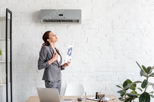 Preview: pretty businesswoman waving newspaper while standing near workplace under air conditioner