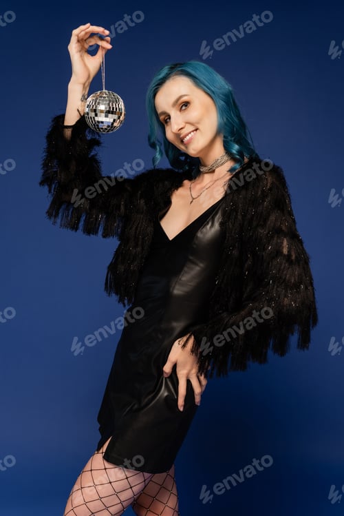 Preview: smiling woman in black faux fur jacket posing with shiny disco ball and hand on hip isolated on blue