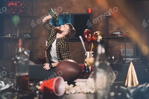 Preview: Bearded young man with hangover drinking wine from bottle after party