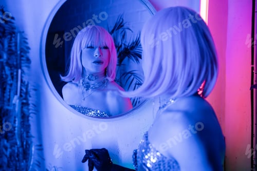 Preview: Blurred drag queen in wig and top looking at mirror in neon light at home