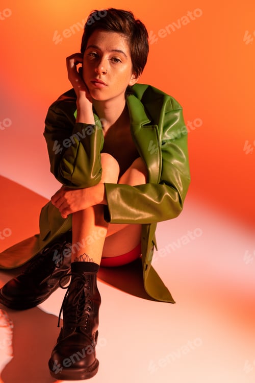 Preview: full length of dreamy woman in green leather jacket and black boots sitting and looking at camera on